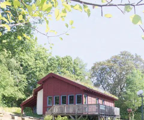 Das rote Naturschutzhaus steht zwischen Bäumen auf einer grünen Anhöhe.