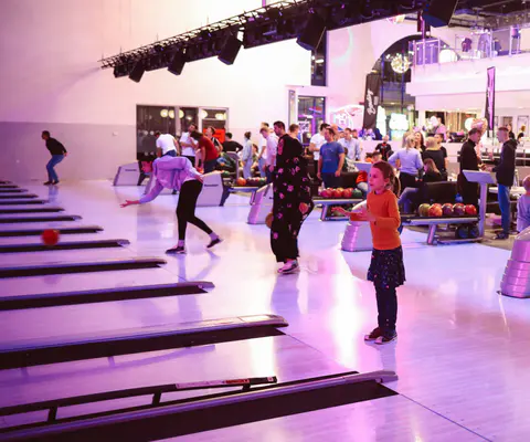 Menschen und Kinder spielen auf mehreren Bowlingbahnen in einer hellen Halle.