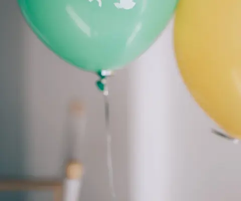 Ein grüner und ein gelber Luftballon hängen vor einer hellen Wand.