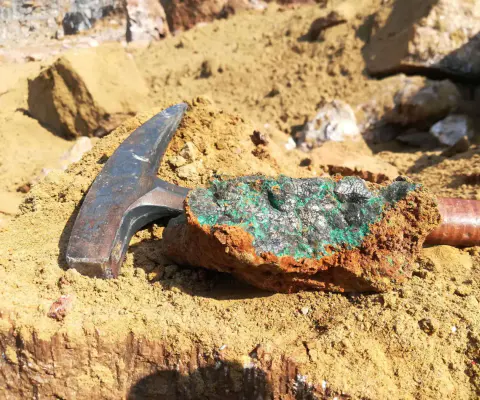 Ein Hammer liegt neben einem Gesteinsbrocken mit grünlichen Mineralspuren im Sand.