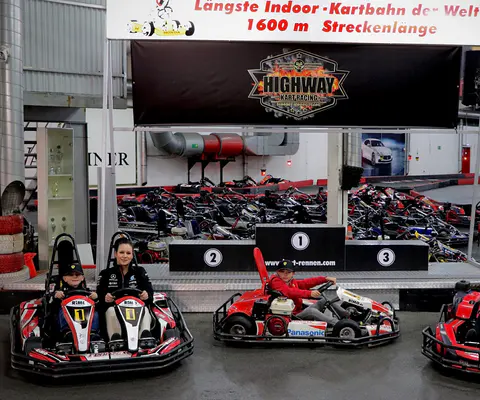 Mehrere Personen stehen in einer Indoor-Kartbahn mit Streckenführung im Hintergrund.