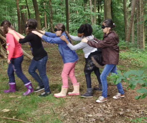 Fünf Kinder mit verbundenen Augen stehen im Wald hintereinander.