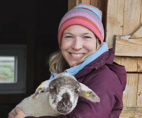 Eine Frau hält ein Lamm vor einer Holzwand im Arm.