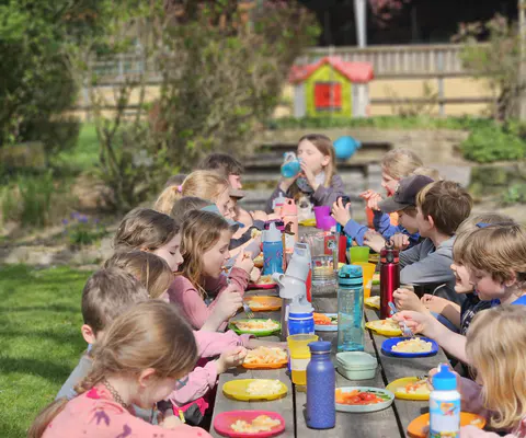 Kinder essen gemeinsam an einem langen Tisch im Garten.