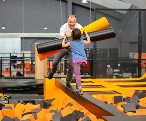 Ein Mann und ein Kind balancieren über eine schmale Plattform im Trampolinpark.