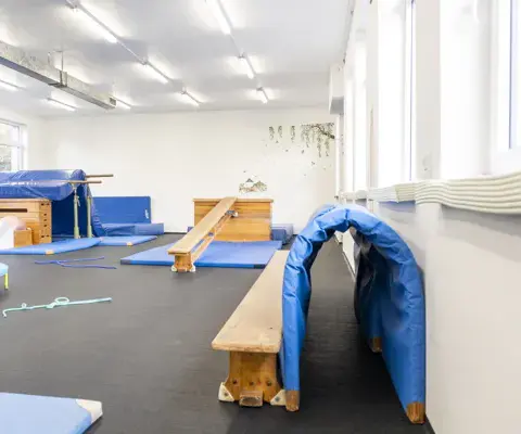 Ein heller Turnraum mit Matten, Bänken, Balanciergeräten und einer kleinen Sprungfläche.