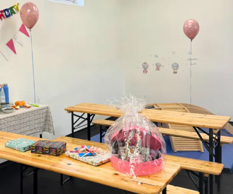 Ein dekorierter Raum mit Geburtstagsbanner, Ballons, Bänken und gedecktem Tisch.